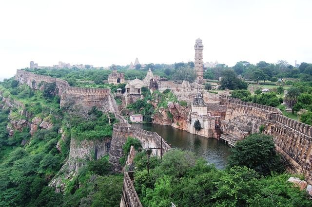 Aerial-style view over Chittorgarh Fort's walls, a water reservoir and greenery, with a tall stone tower on the skyline